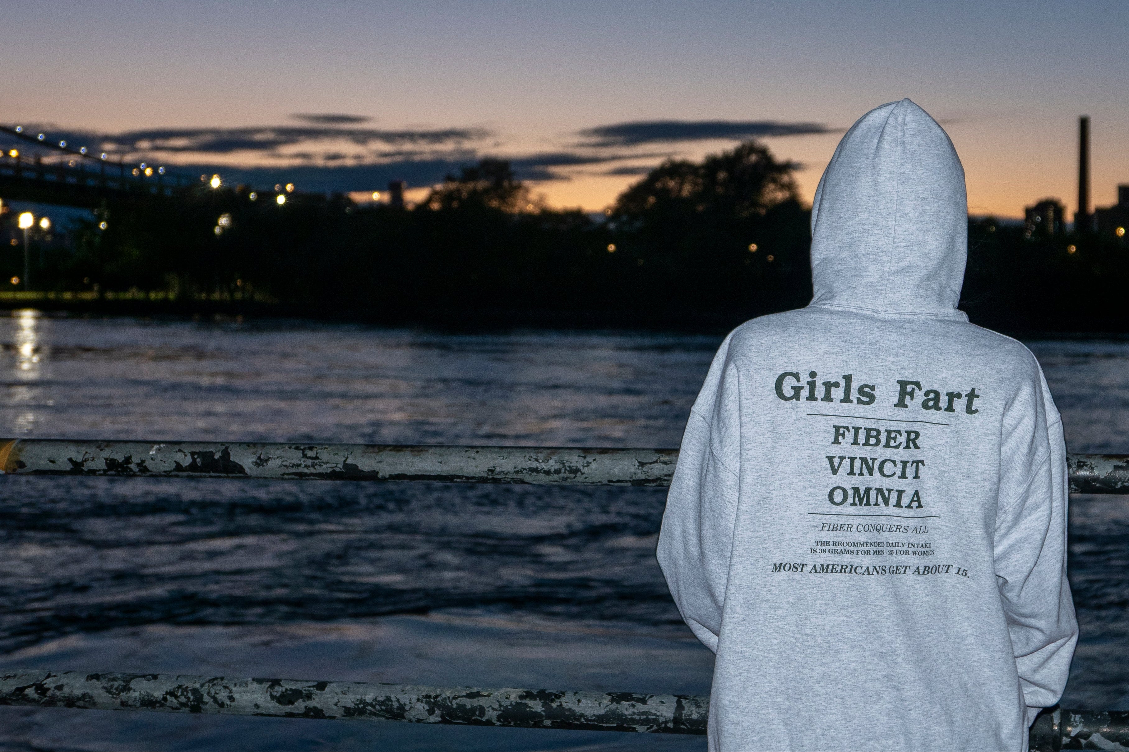 Person wearing a hoodie with text standing by a waterfront at dusk